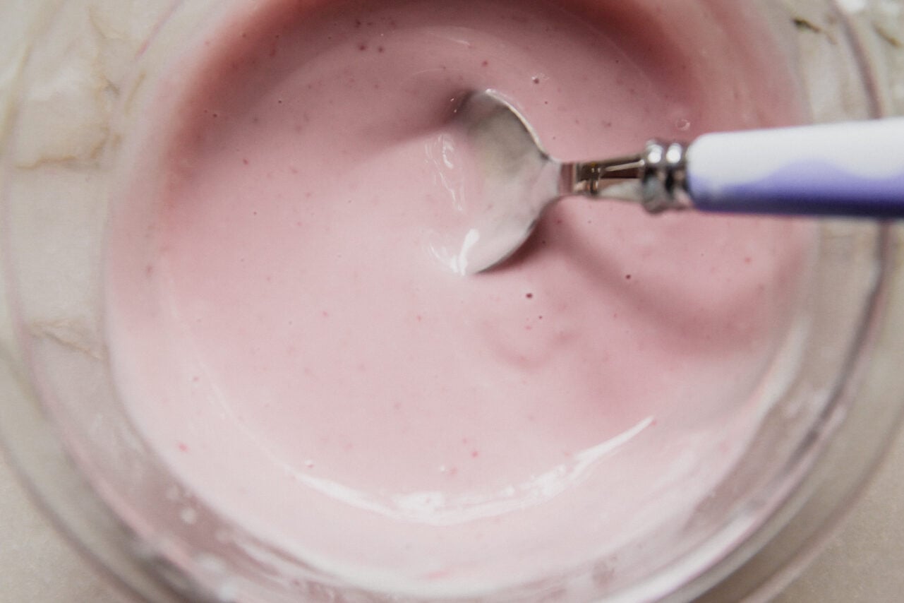 A glass bowl with a yogurt blackberry icing mixture being mixed by a spoon.