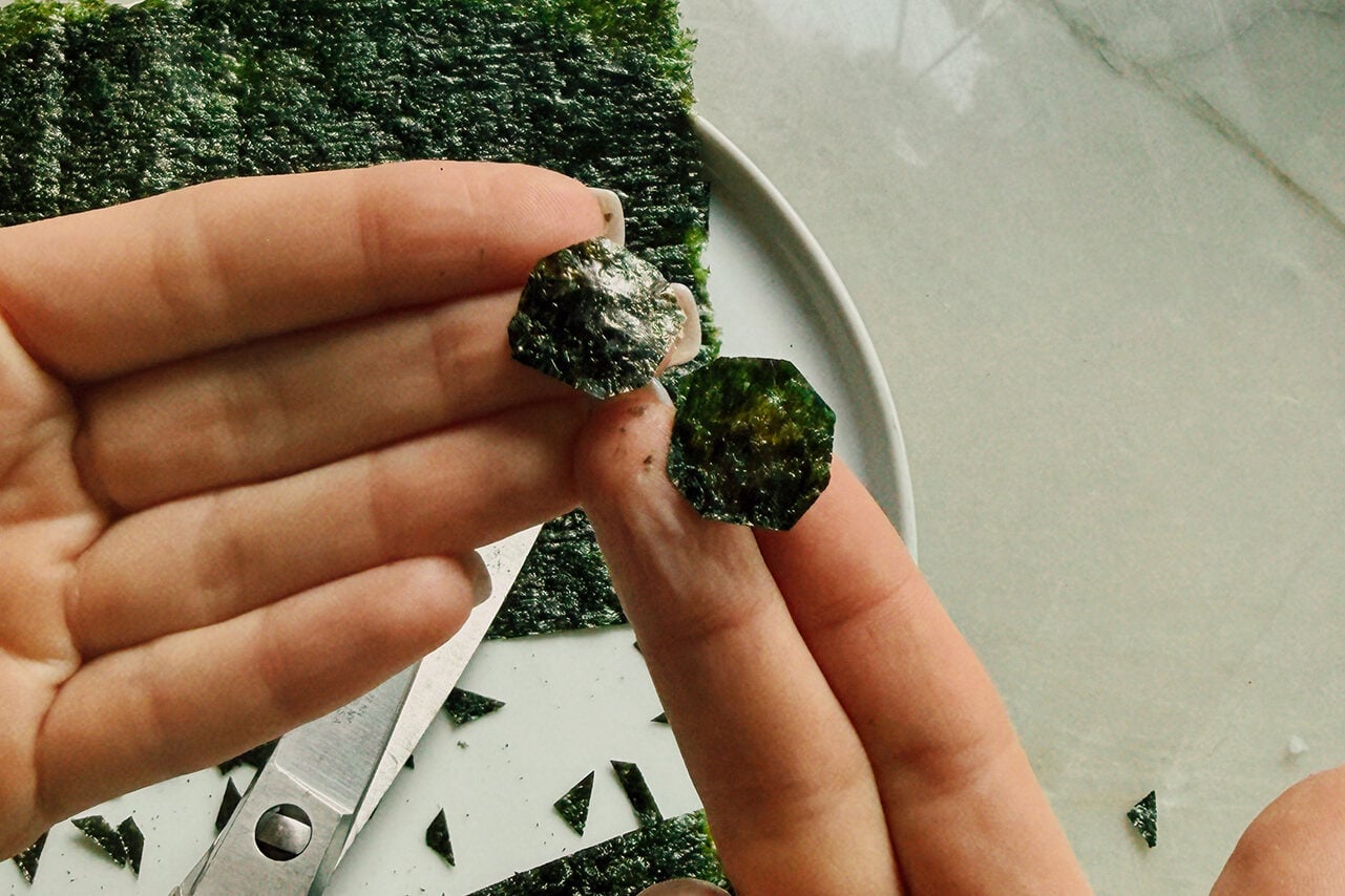 Seaweed cut into soccer balls being held in a hand above scissors and extra seaweed.