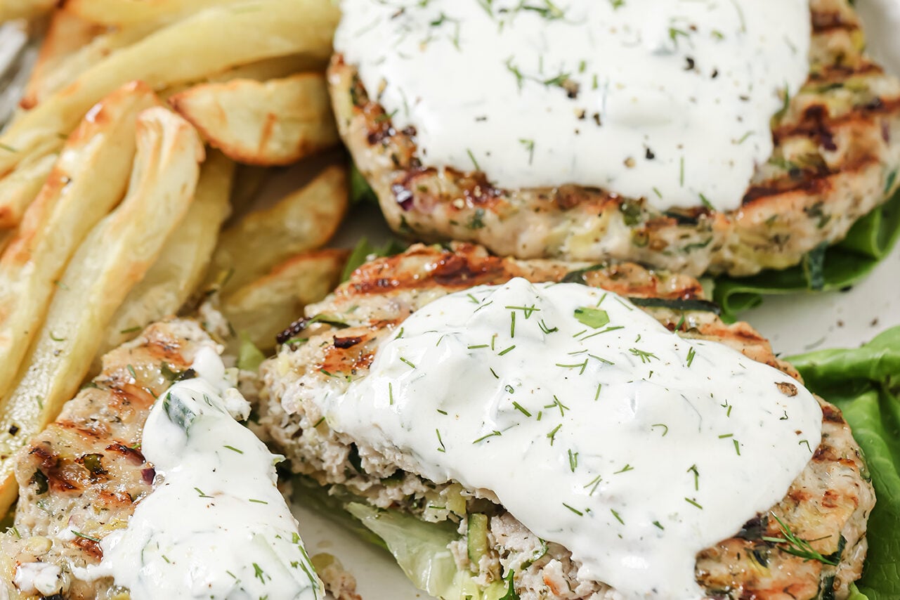 A white plate with Greek chicken burgers on it topped with tzatziki sauce and roasted potatoes next to it.