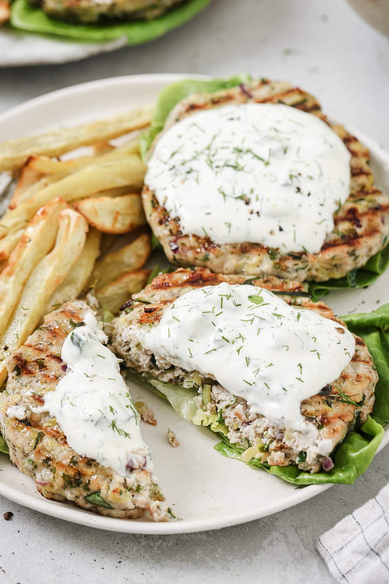 A plate topped with lettuce topped with Greek chicken burgers topped with tzatziki sauce with roasted potato fries next to it.