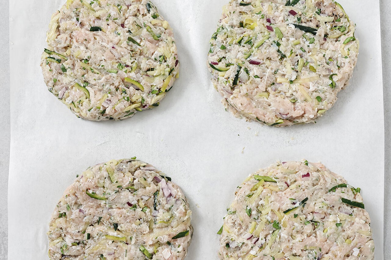 A piece of parchment paper with four Greek chicken burgers formed before cooking.