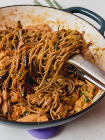 Chicken yakisoba in a large cast iron pan with tongs pulling up the noodles